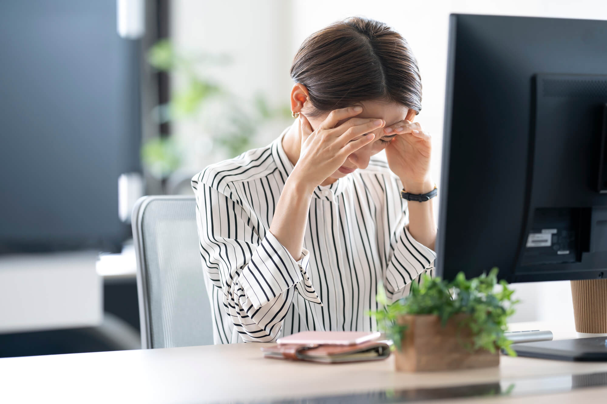 パソコンの前に座った状態でおでこに両手を当てて下を向く女性