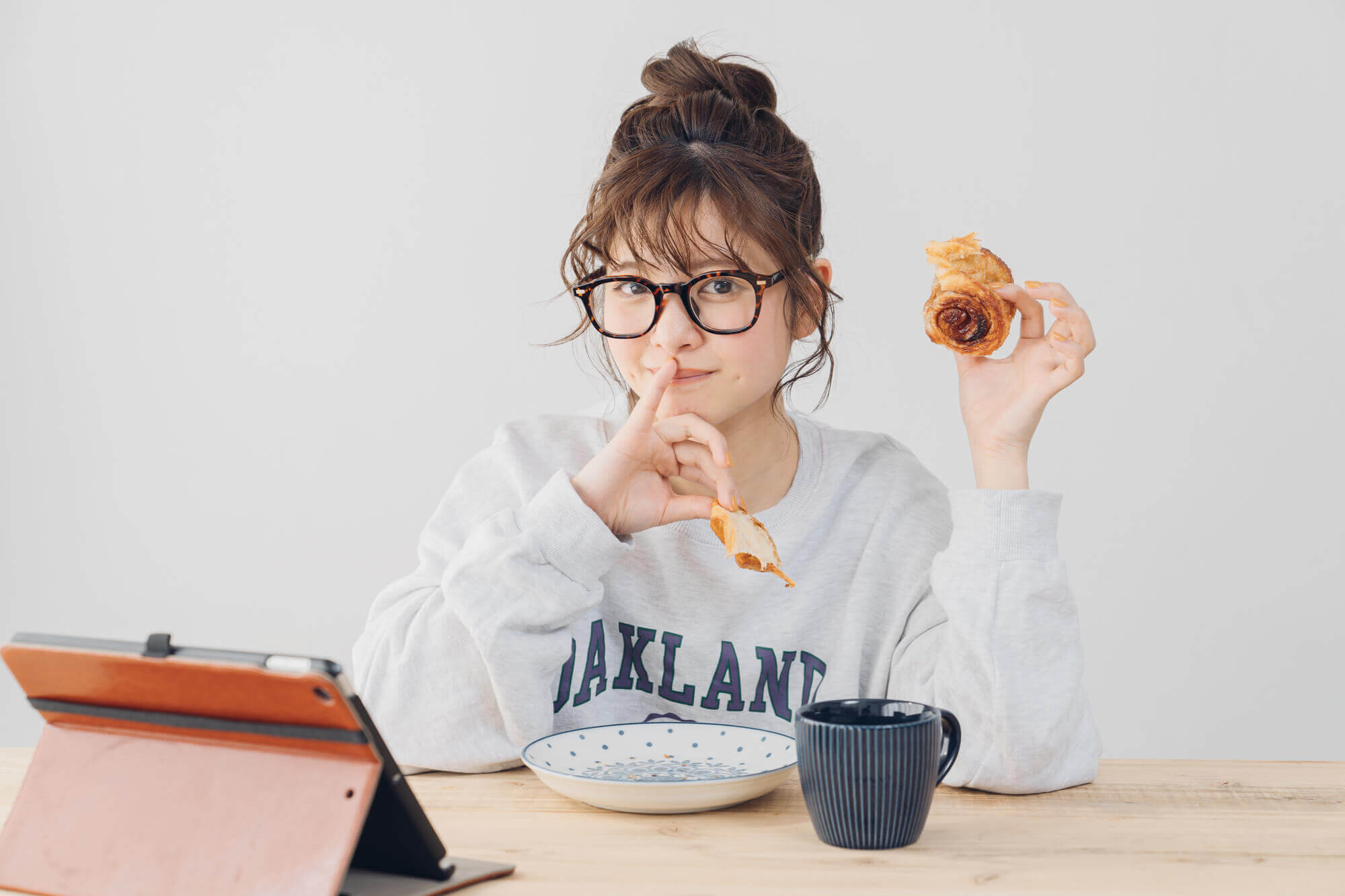 開いたノートパソコン、マグカップ、皿が置かれたテーブルに座り、パンを食べている女性